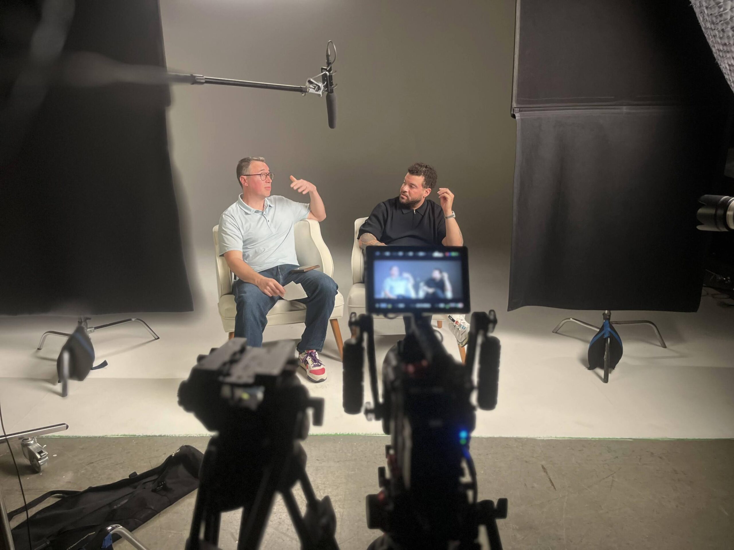 A big brother duo sit in a video interview setup, while a RED camera on a tripod is blurred in the foreground.
