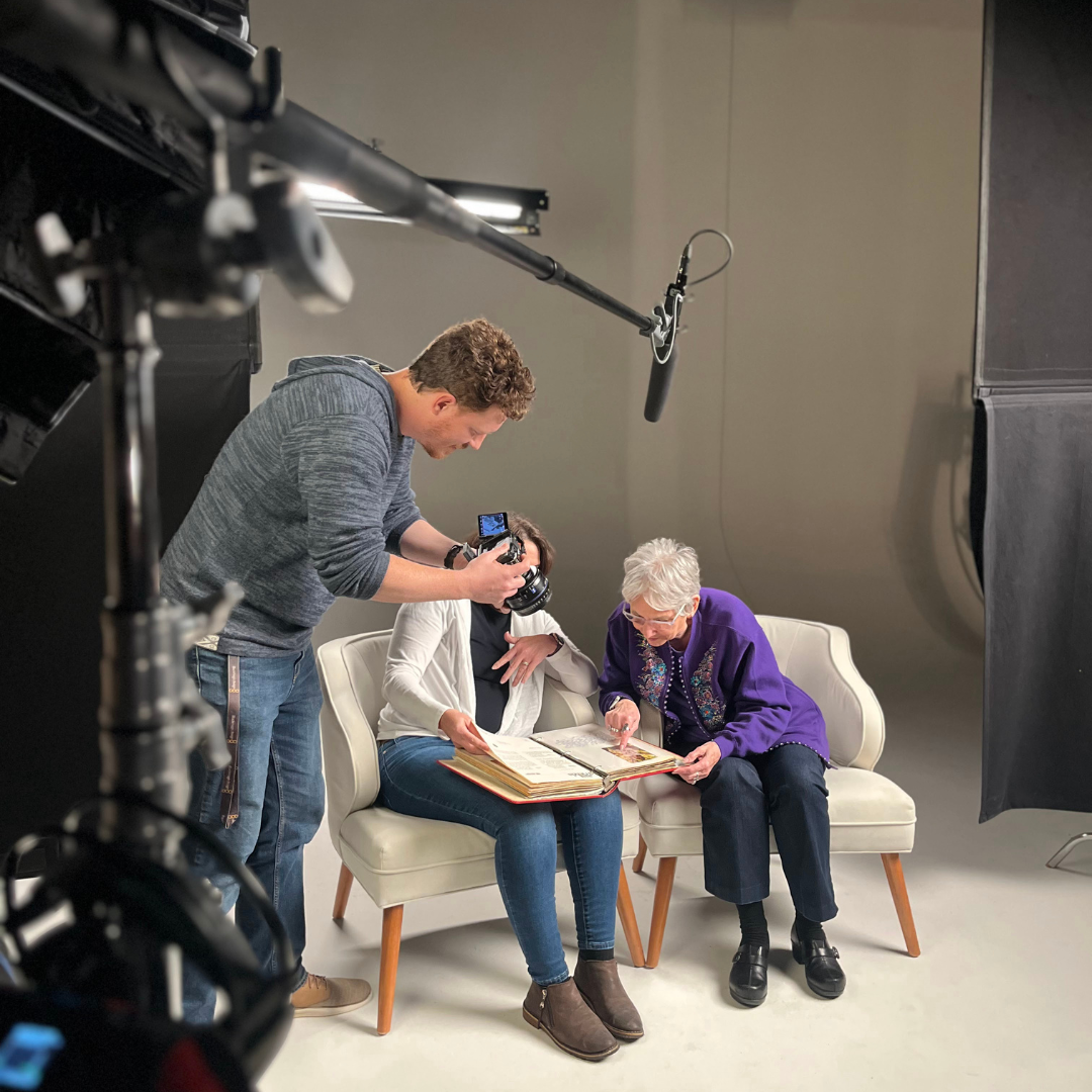 In the studio, two women sit in an interview shoot and look through a photo album together. John, Backflip's director, films from above them where a boom mic is prominently seen.
